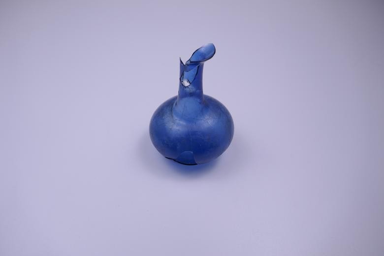 Hier sehen wir ein kleines blaues Glasfläschen, ein sogenanntes Balsamarium auf weißem Hintergrund, welches als Salbgefäß gedient hat. Es hat einen runden, breiten Bauch mit einem schmalen, länglichen Hals. Am Hals fehlt etwas Glas, dort sind Glasscherben abgebrochen.