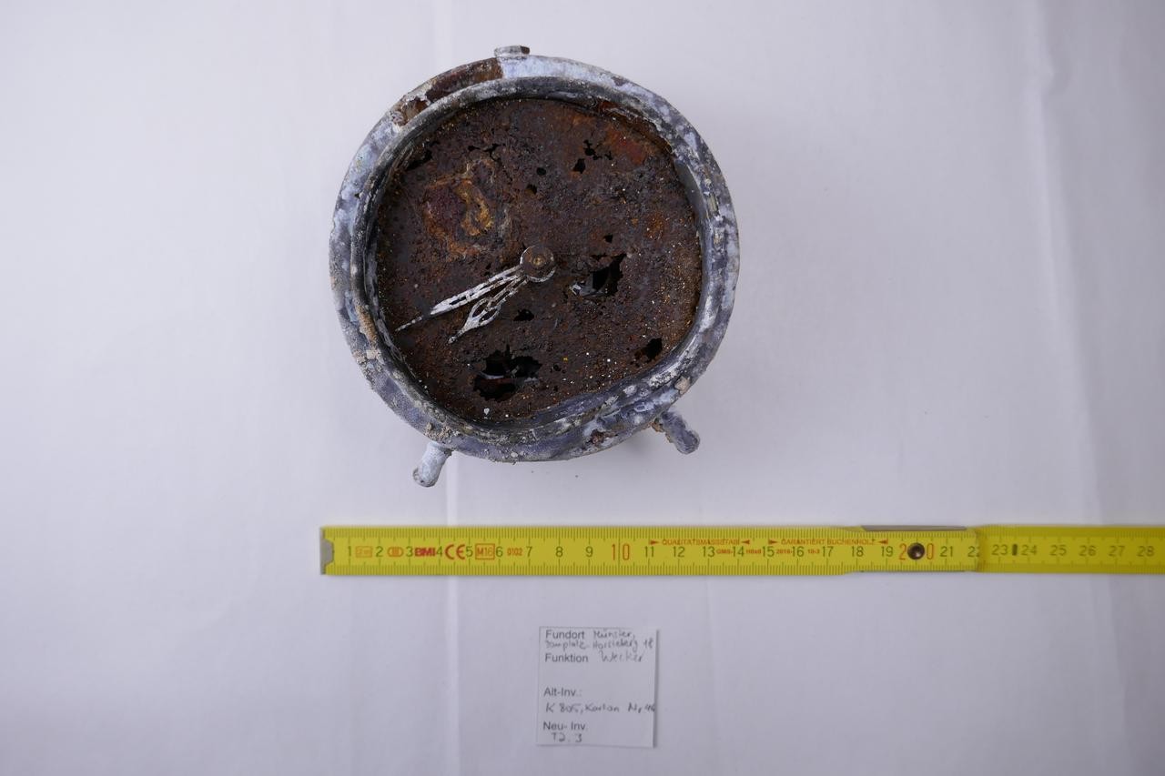 Foto eines durch Bomben beschädigten Weckers aus dem 2. Weltkrieg. Die Zeiger des Weckers sind auf etwa 7:43 Uhr gerichtet. Der Wecker ist in einem schlechtem Zustand und weites gehend verrostet.
