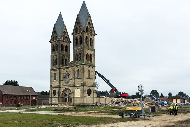 Zu sehen ist die Immerather Kirche, die von Baggern zerstört wird. Drumherum ist alles leer, nur noch ein paar Menschen, wenige Häuser und ein Bagger. Das Wetter ist bewölkt.