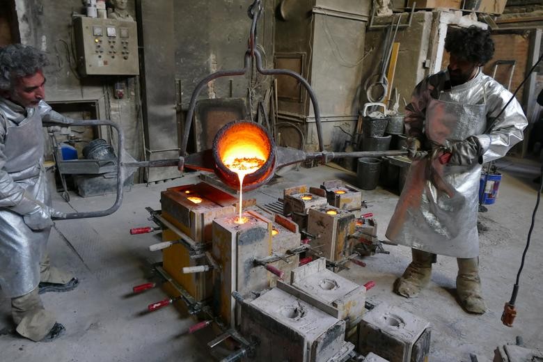 Ein Foto von zwei Männern, die flüssige Bronze in eine Gussform gießen. Sie tragen silberne Schutzkleidung. Sie gießen aus einem Eimer, den sie mithilfe von zwei Stangen um ihn zu drehen. Aus dem Eimer läuft das flüssige Metall in eine Gussform in Form eines aufrechten Quaders mit einem Loch an der oberen Seite. Im Hintergrund sind weitere Gussformen zu sehen.