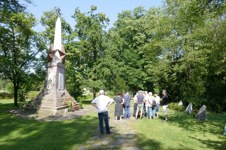 Eine Gruppe Menschen steht vor dem sowjetischen Denkmal auf dem Friedhof des Stalag 326 umgeben von Bäumen, durch dessen Baumkronen die Sonne durchscheint.