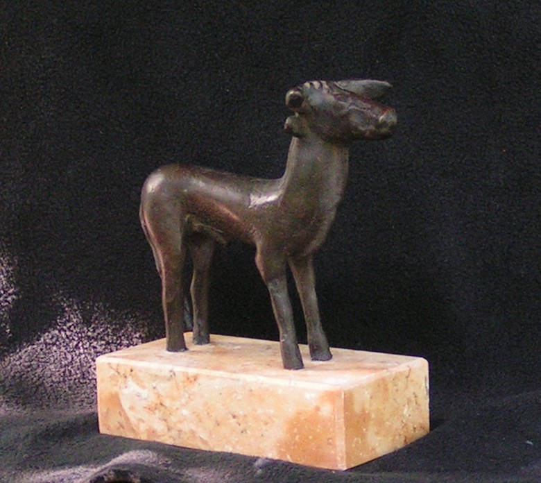 Hier sieht man einen dünnen jungen Stier aus Bronze. Seine Oberfläche ist glatt. Er hat eine Nase, einen langen Schwanz, und ein Horn auf der linken Seite. Das rechte ist abgebrochen. Seine Augen sind leicht eingraviert.  An der rechten Seite rage noch so etwas wie Ohren aus seinem Hals / Kopf. Man kann den Übergang von Beinen in die Hufen erkennen.
