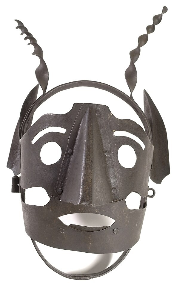 Es ist eine Eiserne Schandmaske auf einem Weißen Hintergrund zu sehen. Man kann deutlich die Form eines Gesichtes erkennen.