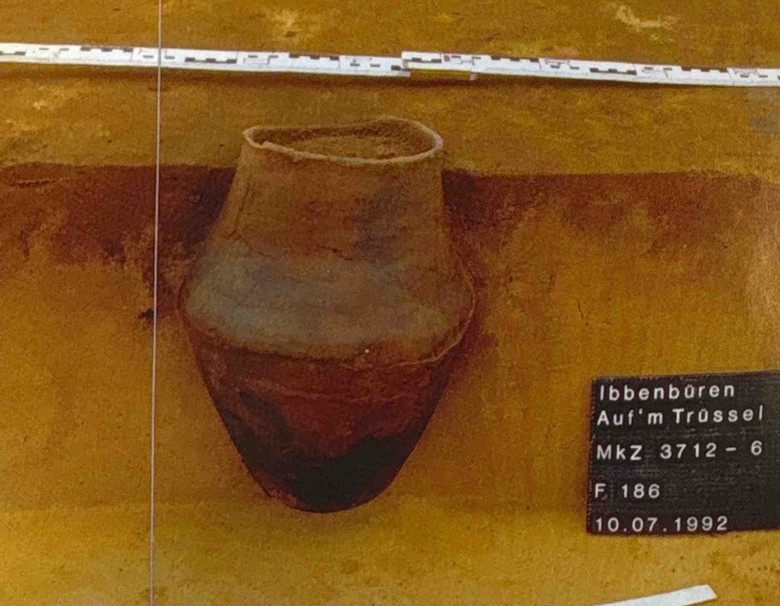 Eine bräunliche Urne im Profil des Befunds, der die lehmige Erde dunkelbraun färbt, rechts ist eine Fototafel, auf der „Ibbenbüren Auf’m Trüssel“ zu lesen ist.