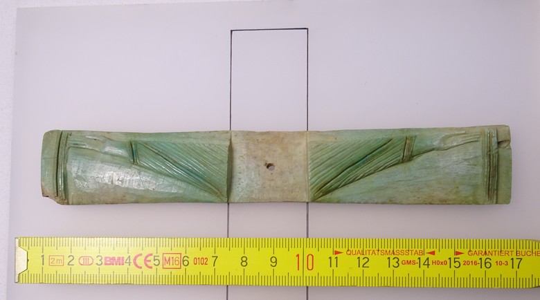 Fotographie des Querbalkens des aus Knochen gemachten Altarkreuzes vor einem weißen Hintergrund. Es besitzt eine grünliche Farbe. Zur Referenz wurde ein Zollstock hinzugelegt. Dieser zeit, dass der Balken ca. 16 cm lang ist. Auf beiden Seiten sind die Arme Christi zu sehen, während der senkrechte Balken nur als Umriss dargestellt werden kann.