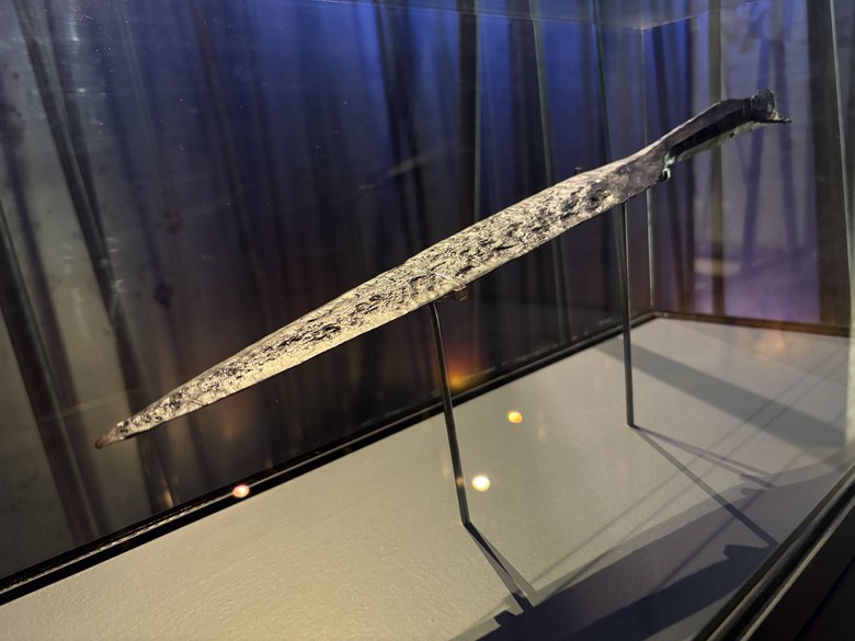 Auf dem Bild sieht man eine längliche, etwas verrostete Lanzenspitze, die von Oben beleuchtet wird und am unteren Teil Verzierungen besitzt.