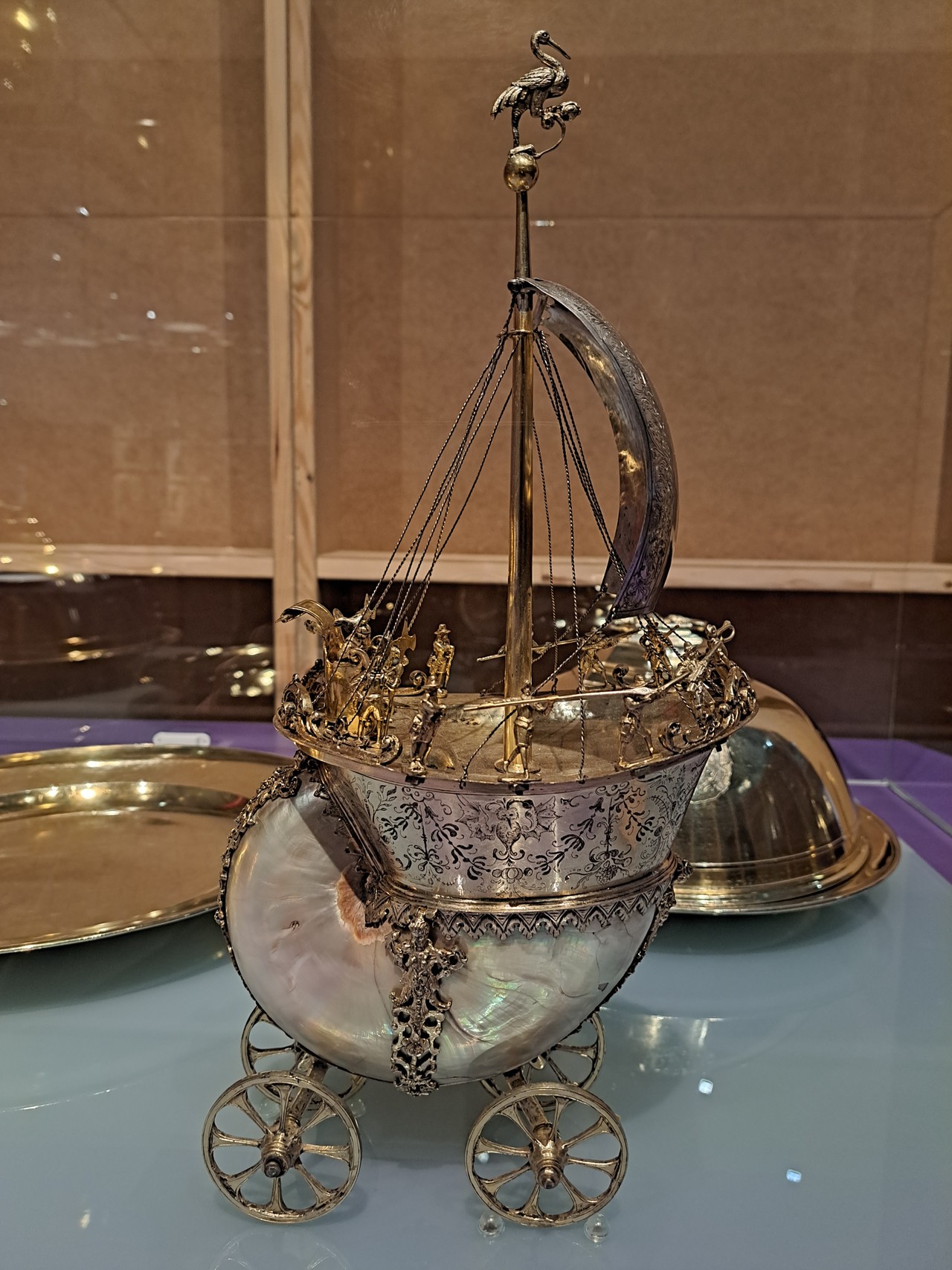 Das Tafelschiff ist von der Seite zu sehen und steht mittig im Bild. Es reicht von der unteren Bildkante bis zur oberen. Der obere Teil des Tafelschiffs sieht aus wie ein Schiff, nur dass der Schiffskörper oval ist und an den Seiten mit schwarzen Blumenranken verziert ist, die symmetrisch sind. In der Mitte der Verzierungen sieht man eine Mischung aus Vogel und Fledermaus. Es hat Federn und einen langen Schnabel, aber die Flügel sehen aus wie bei einer Fledermaus. Außerdem ist der Schiffskörper an den Seiten silbern. Das Deck ist golden und konkav, also zur Mitte hin nach innen gewölbt. Genau an dieser Stelle ragt der Mast, welcher auch golden ist, in die Höhe. Das Ende des Mastes bildet eine goldene Kugel, auf der ein goldener Kranich sitzt. Der Kranich schaut nach rechts, ist also ebenfalls von der Seite zu sehen. Mit seiner linken Kralle steht er auf der Kugel und mit seiner rechten Kralle hält er einen goldenen Stein, welcher wiederum durch eine goldene Verbindung mit der Kugel verbunden ist. Der Hals des Kranichs ist s-förmig nach hinten gebogen, sodass der Schnabel nicht über den Körper hinausragt. Das viereckige Segel ist silbern und nach rechts gewölbt, also von der Seite zu sehen. Es ragt nicht über das Schiff hinaus. Man kann noch leicht eingeritzte Verzierungen erahnen. Das Segel ist kurz unter der Kugel am Mast befestigt. Die untere Seite wird mit feinen silbernen Ketten gehalten. Sie verlaufen schräg nach links und sind an der Kante des Decks befestigt. Vom Mast, kurz unter dem Segel, gehen weitere solcher Ketten nach links und rechts ab und sind überall rund um das Schiff befestigt. Diese Ketten sind aber, im Gegensatz zu denen, die das Segel halten, nicht stramm. Vorne und hinten an den Schiffskanten gibt es goldene Verzierungen. Die Seiten sind leer. Daher kann man die Verzierungen nicht ganz sehen. Auf der linken Seite des Schiffs steht ein goldener Thron, den man auch von der Seite sieht. Die Lehne ist nach hinten gewölbt und bildet am Ende eine Ro