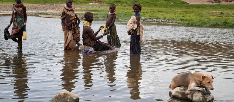 Auf diesem Bild sind fünf Kinder und ein Hund zu sehen, die sich in einem Fluss befinden. Die Kinder gehören dem Turkana Stamm aus Nordkenia an und tragen traditionelle, bunte, Kleidung. Ein Kind sitzt auf einem Stein im Fluss, die anderen stehen in der Nähe im Fluss. Ein Hund liegt auf einem Stein in der Nähe der Kinder im Fluss.