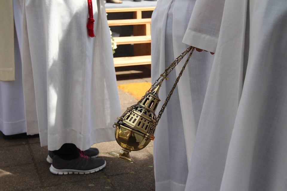 Das Bild ist ein Farbphoto. Es zeigt ein Weihrauchgefäß, dass von Messdiener:innen geschwenkt wird. Von diesen ist jedoch nur der untere Teil des Gewands erkennbar. Das Weihrauchgefäß ist golden und verziert.