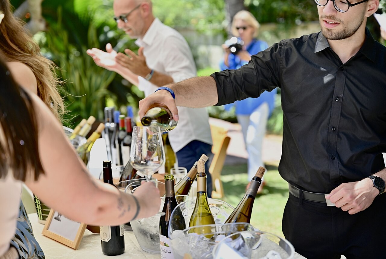 Fotografie eines Mannes, der draußen an einer Weinbar einer Frau ein Glas Wein einschenkt. Der Fokus liegt auf dem Glas, der Flasche und den Händen.