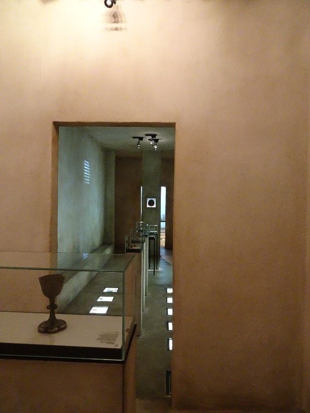 Das Bild ist ein Farbphoto. Es zeigt den Kubus, in dem das Exponat liegt, von innen. Im Vordergrund erkennt man eine Vitrine, in der ein Messkelch ausgestellt ist. Dahinter befindet sich ein Durchgang in einen weiteren Raum des Kubus‘. Dort befinden sich weitere Vitrinen und beleuchtete Einlassungen im Boden für weitere Exponate. An der linken Wand ist eine Projektion erkennbar. Die Wände wirken wie aus altem, mit Mörtel verputztem Stein.