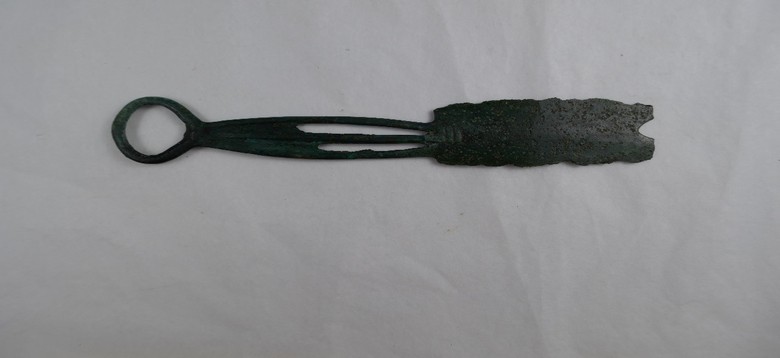 Foto eines zweischneidigen Rasiermessers aus der späten Bronzezeit, aufgenommen auf weißem Hintergrund. Links ist der ringförmige Griff mit drei durchbrochenen Stegen zu sehen, rechts die stark korrodierte Klinge mit erkennbar V-förmigem Einschnitt an der oberen Kante. Das Objekt ist vollständig sichtbar und liegt flach, sodass Form und Material gut erkennbar sind.